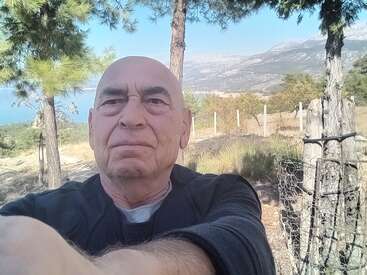 Un homme âgé prend un selfie en plein air, debout près d'arbres, avec des montagnes, une clôture et un ciel bleu clair en arrière-plan, profitant d'un paysage paisible.