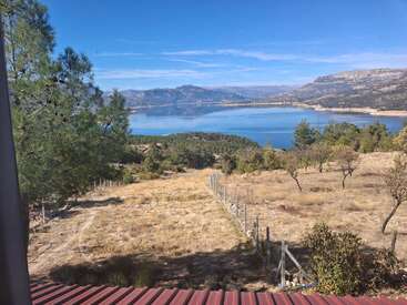 A serene landscape features a clear blue lake surrounded by mountains, pine trees, and dry grass. A metal fence runs through the foreground under a blue sky.