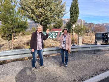 Deux personnes souriantes saluent l'appareil photo au bord d'une route rurale, entourée d'arbres et de montagnes. Un panneau vert et une camionnette garée sont visibles à proximité.