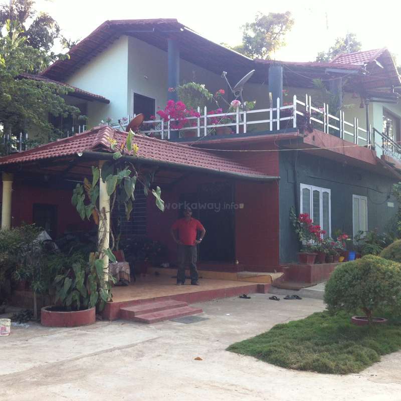 The image depicts a two-story house with a red roof, featuring a man standing on the porch and a balcony adorned with plants and flowers.