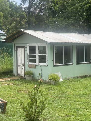 A small, mint-green house with white windows and door, surrounded by green grass, potted plants, and trees. Sunlight filters through, creating a peaceful, rustic atmosphere.