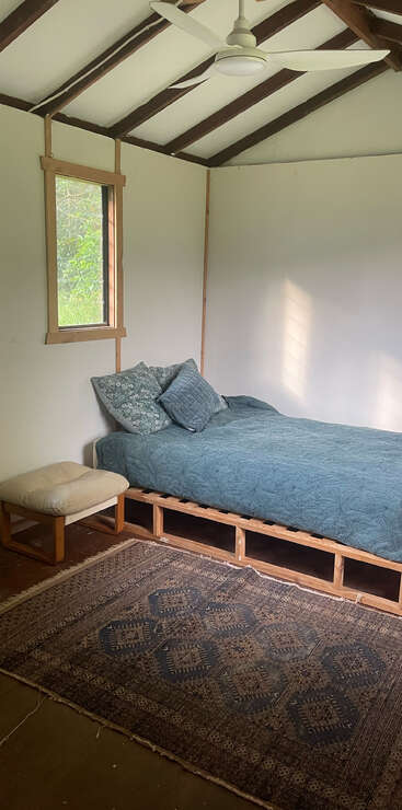 This simple room features a wooden bed with blue bedding, a rug, a cushioned stool, a window, white walls, and a ceiling fan.