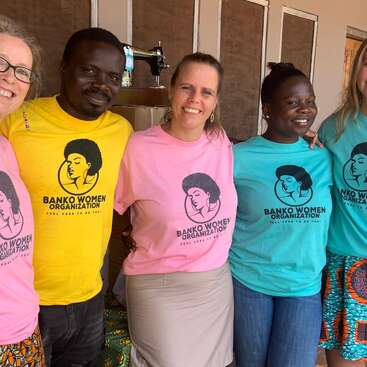 A group of smiling people wearing colorful "Banko Women Organization" shirts stands together, arms around each other, promoting unity, empowerment, and support for women's causes.