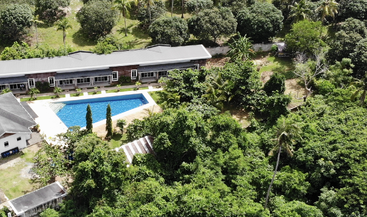 A modern building with a gray roof sits beside a rectangular swimming pool, surrounded by lush green trees and dense forest, creating a tranquil retreat.