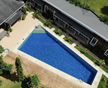 This image shows a rectangular blue swimming pool surrounded by a tiled deck, greenery, some lounge chairs, and buildings, viewed from above on a sunny day.