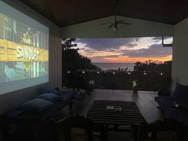 L'espace de vie extérieur comprend une installation de cinéma confortable avec "The Sandlot" projeté sur le mur, des sièges confortables et une vue imprenable sur l'océan au coucher du soleil.