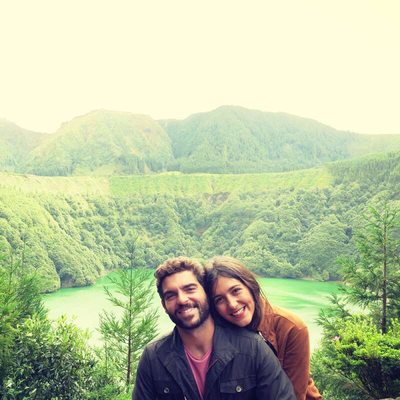 Das Bild zeigt eine ruhige Szene mit einem Mann und einer Frau, die vor einer üppig grünen Bergkette und einem See, umgeben von Bäumen, posieren.