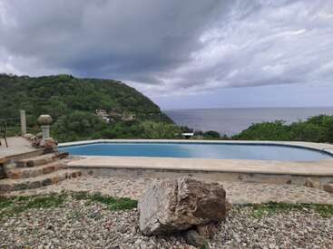 An infinity pool overlooks lush green hills and a calm sea under a dramatic cloudy sky. Stone steps and a large rock complete the peaceful scene.
