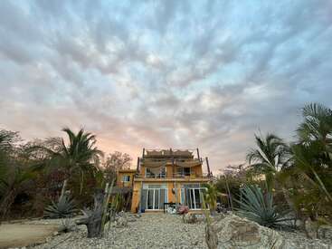 A two-story yellow house with large windows stands beneath a dramatic, cloudy sky, surrounded by palm trees and desert plants, creating a serene tropical atmosphere.