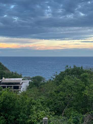 A beautiful landscape shows lush green trees, a building partially hidden, deep blue ocean waves, and a dramatic cloudy sky tinged with soft orange light at sunset.