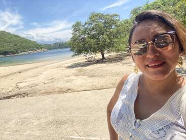 A woman in sunglasses and a white outfit smiles at the camera, standing on a beach with clear blue water, trees, and green hills in the background.