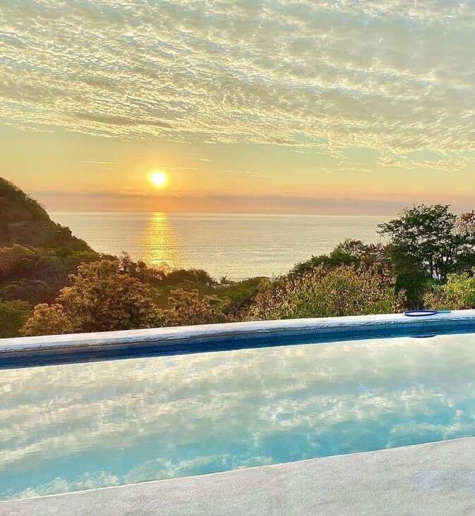 A serene sunset over the ocean is beautifully reflected in a clear swimming pool, surrounded by lush greenery and a peaceful sky filled with soft clouds.