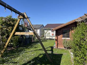 Une scène ensoleillée dans un jardin à l'arrière d'une maison, avec une balançoire en bois, de l'herbe verte, des fleurs blanches éparpillées, un petit abri en bois, des buissons et un ciel bleu au-dessus. Une atmosphère paisible.