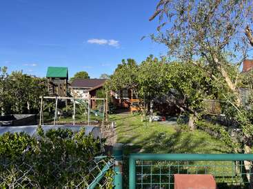 Un jardín soleado en el patio trasero con césped verde y frondoso, árboles, una estructura de madera para jugar, cobertizos y una zona de descanso, rodeado por una valla bajo un cielo azul claro.