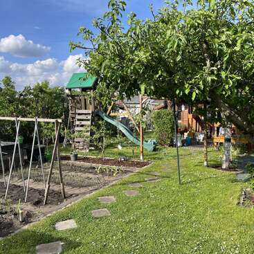 Cette image montre un jardin ensoleillé avec un potager, de l'herbe verte, des pas japonais, une aire de jeux en bois équipée d'un toboggan, des arbres et une maison en bois chaleureuse.