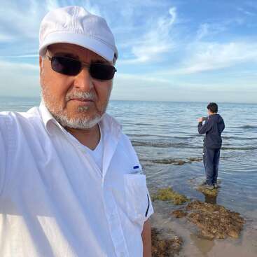 Um homem de óculos escuros e boné branco tira uma selfie na praia, enquanto outra pessoa fica na água de frente para o mar, sob um céu azul.