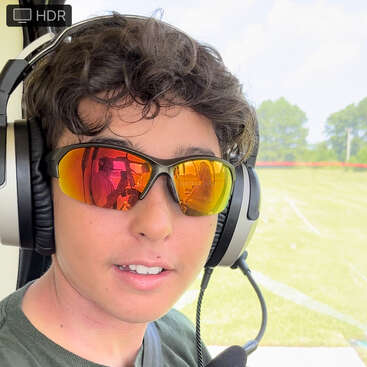 Une personne portant des lunettes de soleil à verres réfléchissants et un casque audio est assise à l'intérieur d'un véhicule, peut-être un hélicoptère. À travers la vitre, on aperçoit un paysage verdoyant et ensoleillé.