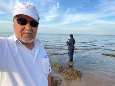 Um senhor de branco tira uma selfie na praia, usando óculos escuros e um boné. Uma pessoa mais jovem está na beira da água, de frente para o mar.