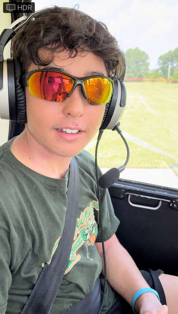 A person wearing reflective sunglasses and a headset sits inside a vehicle, possibly a helicopter. The outdoor scenery is green and sunny, visible through the window.