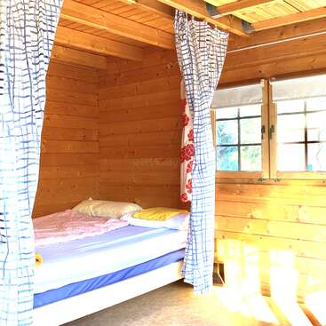 Um aconchegante quarto em uma cabana de madeira, com a luz do sol entrando pela janela, com uma cama com roupa de cama colorida, almofadas e cortinas estampadas para garantir a privacidade.