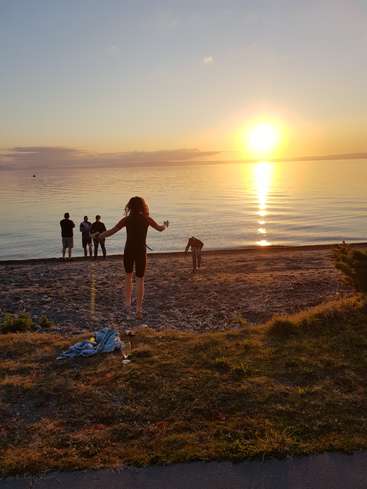 L'image représente une scène de plage sereine au coucher du soleil, avec une femme en combinaison de plongée, un groupe de personnes sur le rivage et un chien, le soleil se couchant sur l'eau.