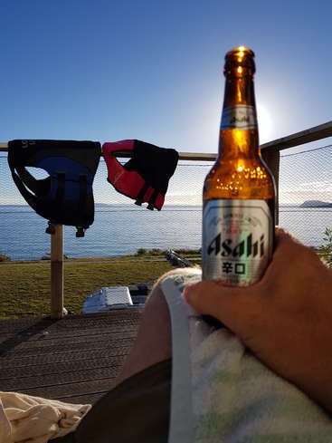 L'image représente une scène sereine d'une personne tenant une bouteille de bière Asahi sur une terrasse surplombant l'océan, avec un gilet de sauvetage accroché à proximité.