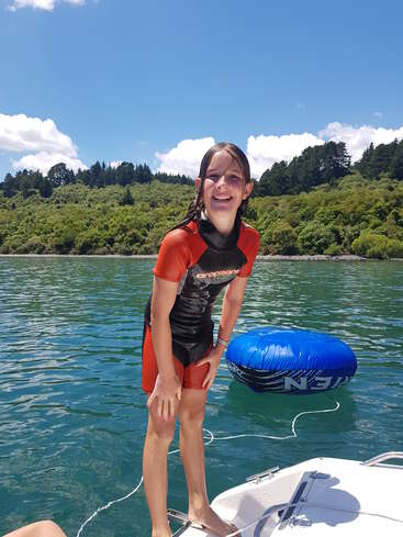 L'image représente une jeune fille debout sur un bateau dans l'eau, vêtue d'une combinaison de plongée noire et orange, avec un tube gonflable bleu derrière elle et une forêt verdoyante à l'arrière-plan.