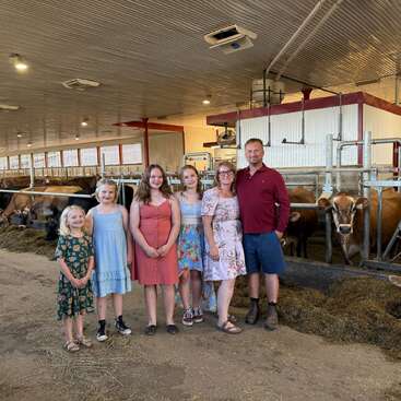 Eine sechsköpfige Familie steht in einem Milchviehstall mit Kühen dahinter und lächelt. Der Stall ist gut beleuchtet und sauber, und alle sehen glücklich aus.