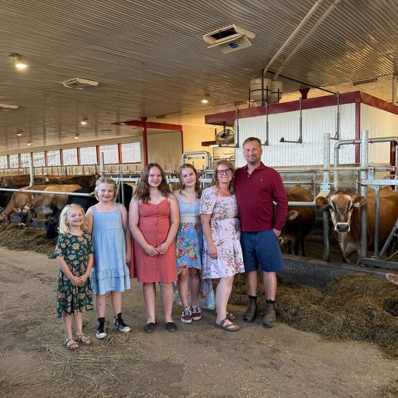 Uma família de seis pessoas está em um celeiro de gado leiteiro com vacas atrás deles, sorrindo. O celeiro é bem iluminado e limpo, e todos parecem felizes.