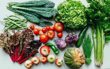Una colorida variedad de verduras y frutas frescas, incluyendo col rizada, lechuga, apio, manzanas, tomates, judías, acelgas, colinabo, calabacín, calabaza y pimientos.