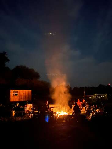 Un grupo de personas se sienta alrededor de una hoguera resplandeciente por la noche, rodeadas de bancos, cabañas rústicas y árboles, con humo elevándose hacia el cielo oscuro.