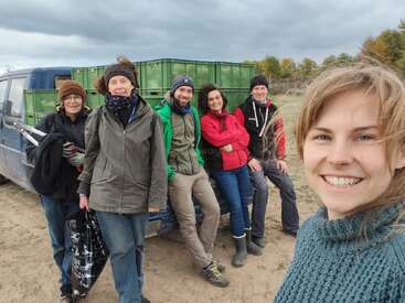 Seis personas posan juntas al aire libre junto a un vehículo con cajas verdes, vestidas con ropa de abrigo para el frío, sonriendo y pareciendo disfrutar de una actividad en grupo en la naturaleza.