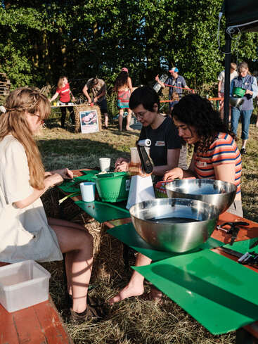 Tres mujeres están sentadas en una mesa al aire libre preparando comida con cuencos y utensilios, mientras otras personas participan en actividades en un entorno soleado, cubierto de césped y con un ambiente festivo.