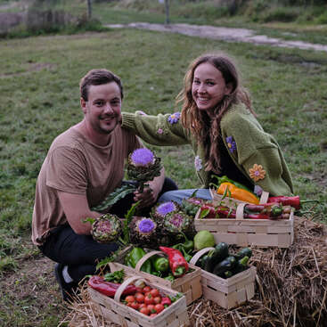 A smiling man and woman pose outdoors with baskets of freshly harvested vegetables and artichokes, sitting on hay in a lush green field, enjoying gardening.