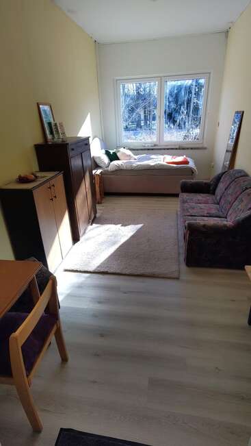Um quarto pequeno e aconchegante com uma cama perto da janela, um sofá com estampa floral, móveis de madeira, um tapete, um espelho na parede e a luz do sol entrando pelas janelas grandes.