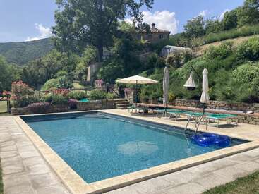 A serene swimming pool surrounded by stone paving, lounge chairs, umbrellas, vibrant flowers, and lush greenery. A charming house sits on the hillside above. Peaceful atmosphere.