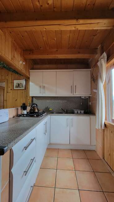 This cozy kitchen features wooden walls and ceiling, white cabinets, a tiled floor, countertop appliances, a window with curtains, and natural sunlight brightening the entire space.