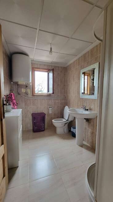 This is a spacious bathroom with beige tiles, a pedestal sink, toilet, mirror, shower, purple laundry basket, cabinet, water heater, and an open window.
