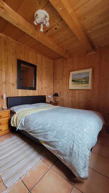 This cozy bedroom features wood-paneled walls, a double bed with a patterned duvet, side tables, lamps, a mirror, framed artwork, and a striped rug.