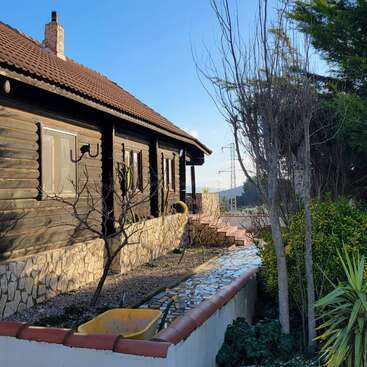 A rustic wooden house with a stone foundation and tiled roof sits beside a small garden, paved path, and yellow wheelbarrow, surrounded by greenery and sunlight.