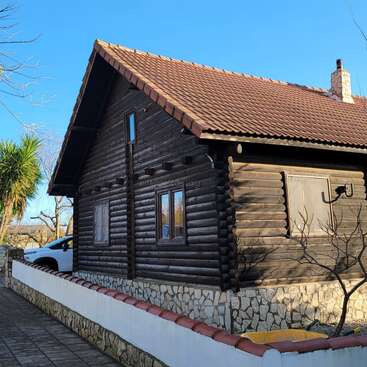 Une cabane rustique en bois avec un toit en tuiles se dresse derrière une clôture de pierres et de murs blancs. Elle est entourée d'arbres sous un ciel bleu clair.