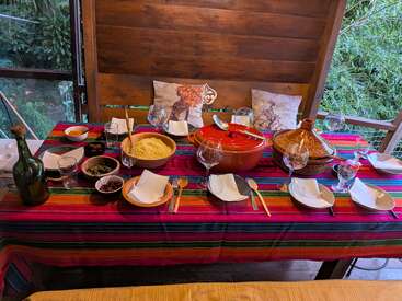 Ein farbenfroher, festlicher Tisch wird im Freien mit bunten gestreiften Tüchern, marokkanischen Tagines, Couscous, Weingläsern, Tellern, Holzutensilien und Kissen mit künstlerischen Tiermotiven gedeckt.