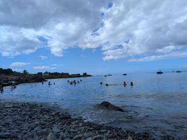 A rocky beach with people swimming, relaxing in calm blue water. Boats float in the distance, under a partly cloudy sky with patches of blue. Peaceful atmosphere.