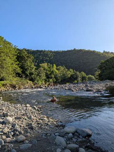 A person swims in a rocky river surrounded by lush green trees and hills under a clear blue sky, creating a peaceful and natural scene.