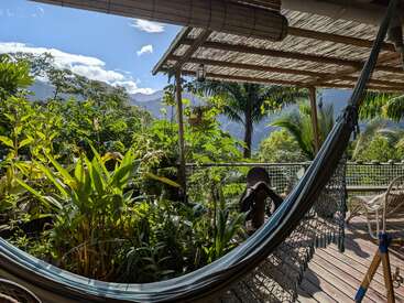 A peaceful porch with a hammock overlooks lush green plants. Wooden decking, mountains, palm trees, and blue skies create a serene tropical retreat in nature.