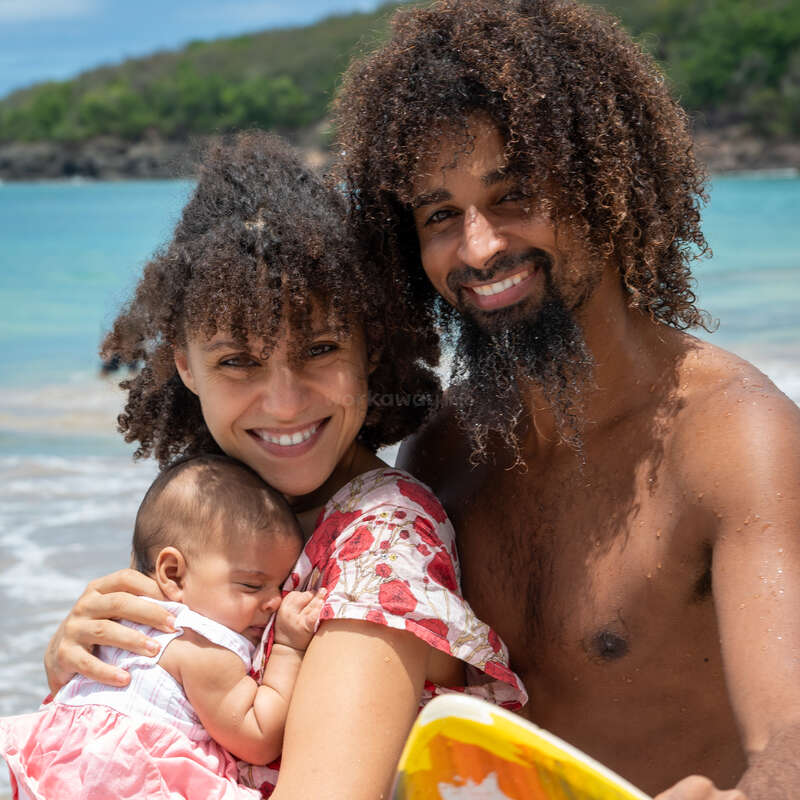 Una familia alegre en la playa. La madre sostiene a un bebé, sonriendo cariñosamente, mientras el padre está de pie cerca, sosteniendo una tabla de surf de colores brillantes. Día soleado.