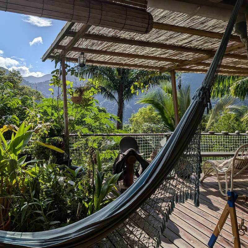 Un porche paisible avec un hamac surplombe des plantes vertes luxuriantes. Les terrasses en bois, les montagnes, les palmiers et le ciel bleu créent une retraite tropicale sereine dans la nature.
