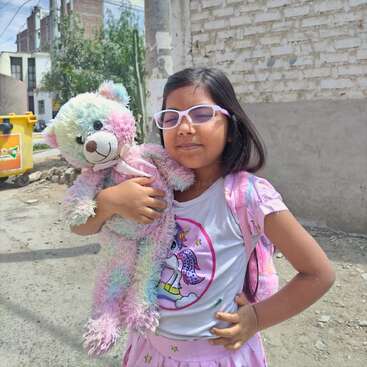 A young girl wearing glasses and a unicorn shirt smiles while holding a colorful teddy bear. She stands outdoors near a brick wall and a yellow bin.