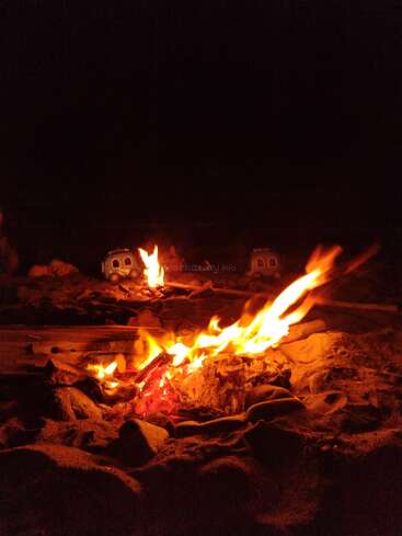A vibrant campfire burns on a sandy beach at night, with glowing flames illuminating the darkness. Two toy vans are visible in the background.