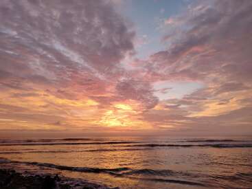 A stunning beach sunset fills the sky with vibrant hues of pink, purple, orange, and yellow, reflecting beautifully on the gentle, rolling ocean waves.